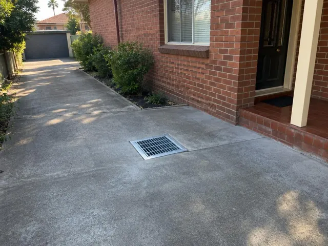 Concrete driveway with a metal drain grate beside a brick house in Rydalmere
